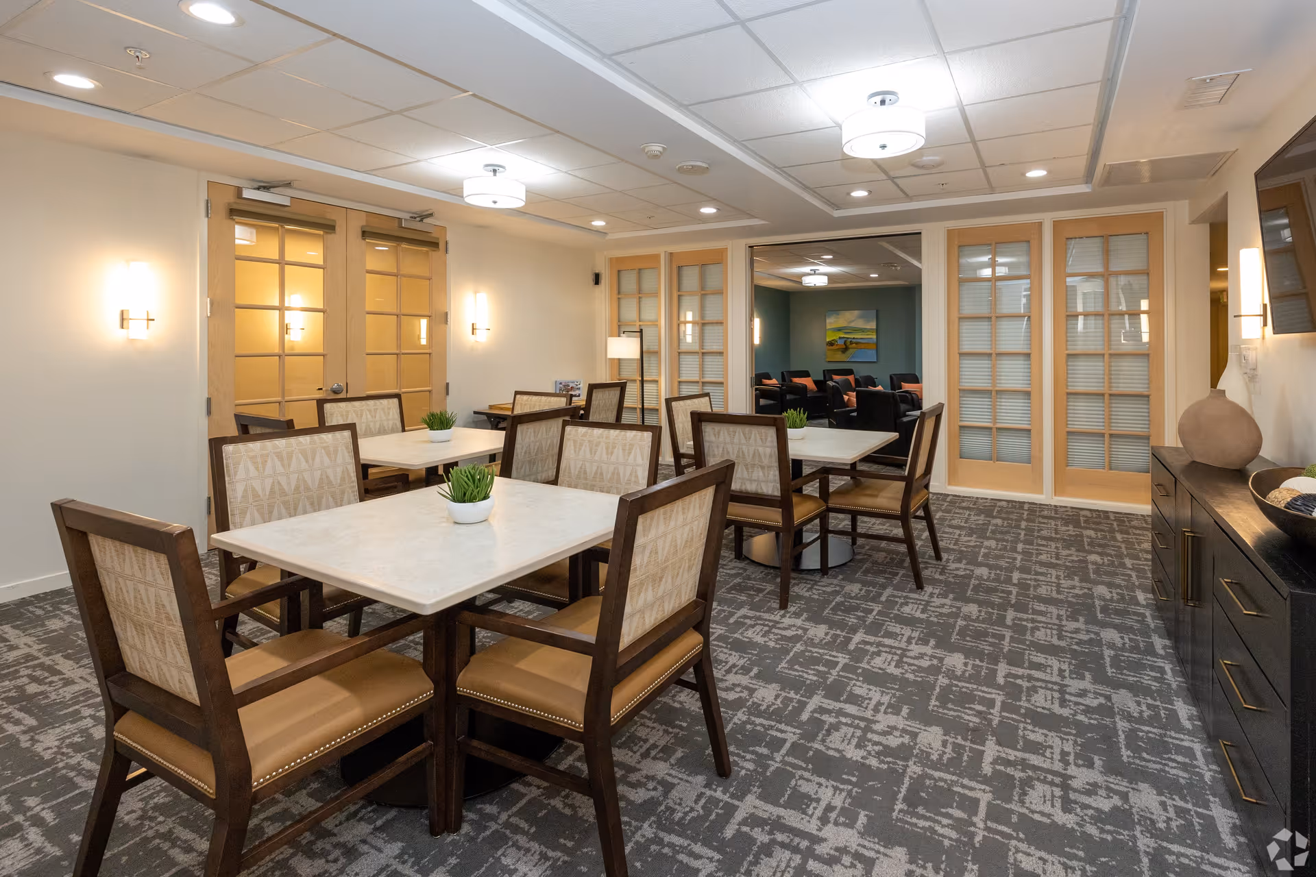 A dining room in a senior living facility with several square tables, each surrounded by four cushioned chairs. The room features patterned carpet, soft wall lighting, and wooden framed glass doors leading to another room with additional seating and artwork on the wall.