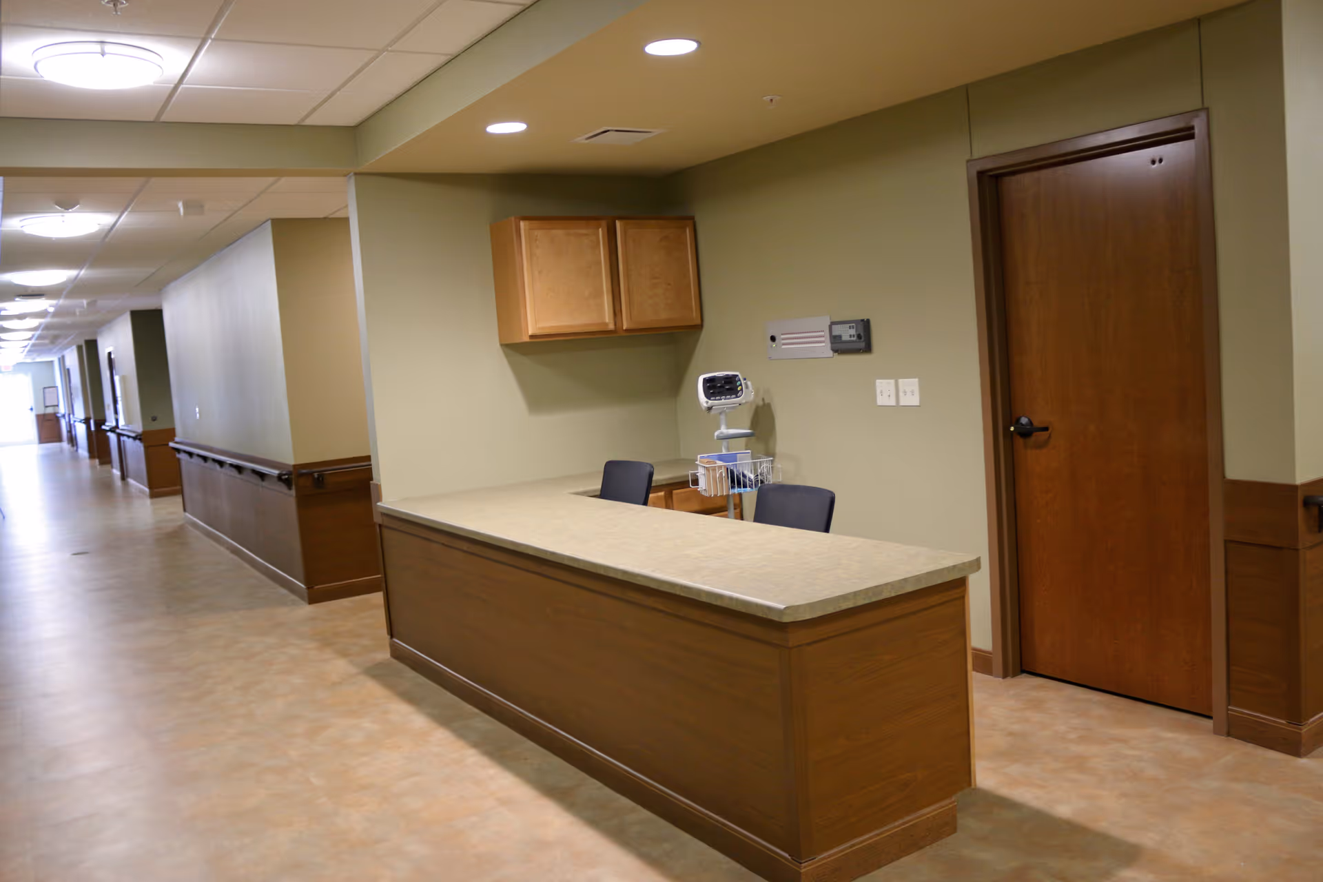 A clean and well-lit hallway in a senior living facility with a wooden reception desk or nurse's station on the right side. The desk has two chairs behind it and a medical monitor on a stand. There are wooden cabinets mounted on the wall above the desk and a closed wooden door next to it. The hallway extends into the distance with handrails along the walls and ceiling lights evenly spaced.