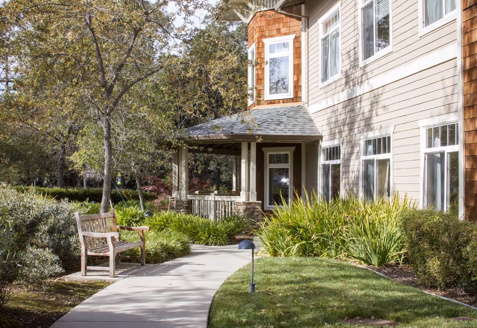 Outdoor garden area at Sunrise of Fair Oaks featuring a curved concrete pathway, a wooden bench, green bushes, trees, and the side of a beige building with multiple windows and a small covered porch.