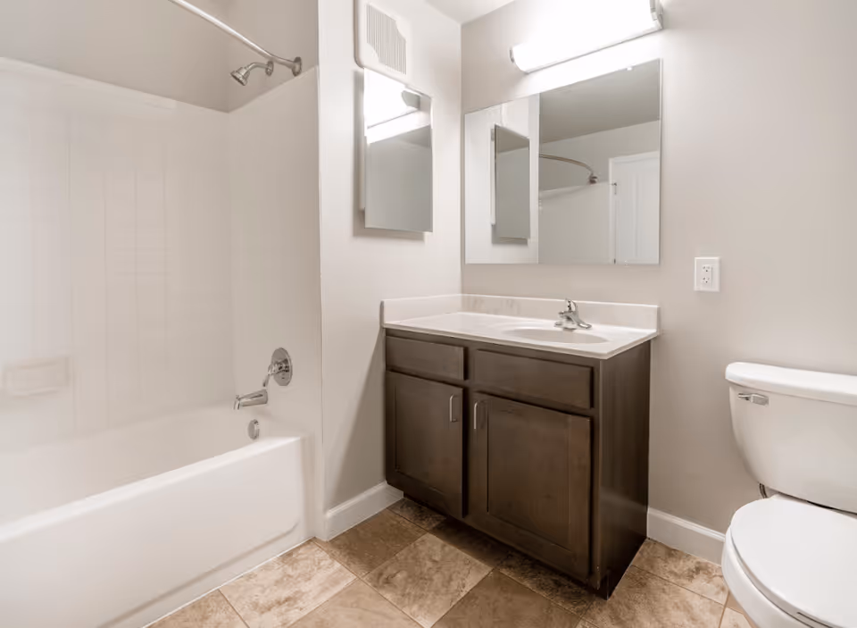 A clean bathroom featuring a white bathtub with a showerhead and a curved shower curtain rod, a dark wood vanity with a white countertop and sink, a large rectangular mirror above the sink, a smaller medicine cabinet mirror on the adjacent wall, a toilet, and beige tiled flooring.