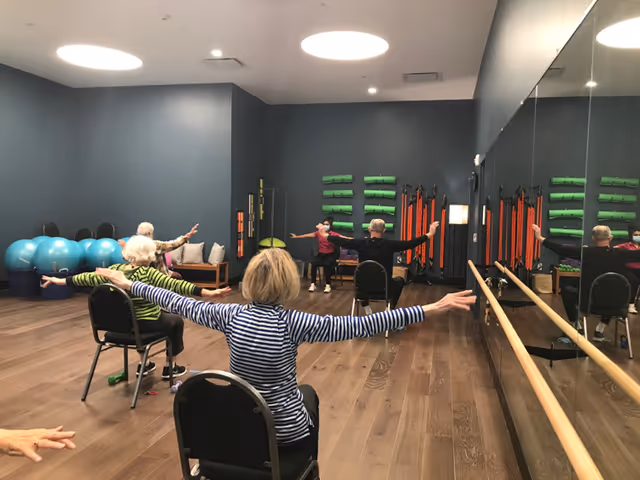 A group of elderly individuals seated on chairs in a fitness room, participating in a seated exercise class with arms extended. The room has wooden floors, a large mirror on one wall, exercise balls, and various fitness equipment mounted on the walls.