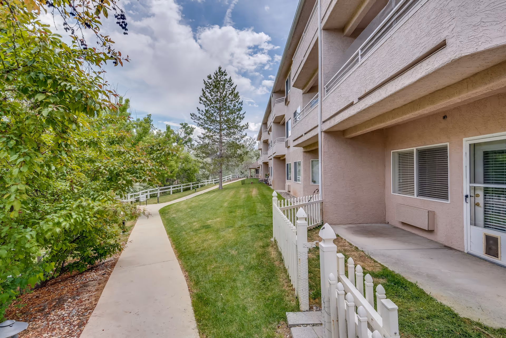 Outdoor view of a senior living facility showing a paved walkway alongside a grassy area with a white picket fence. The building has multiple balconies and windows, and there are trees and bushes along the path under a partly cloudy sky.