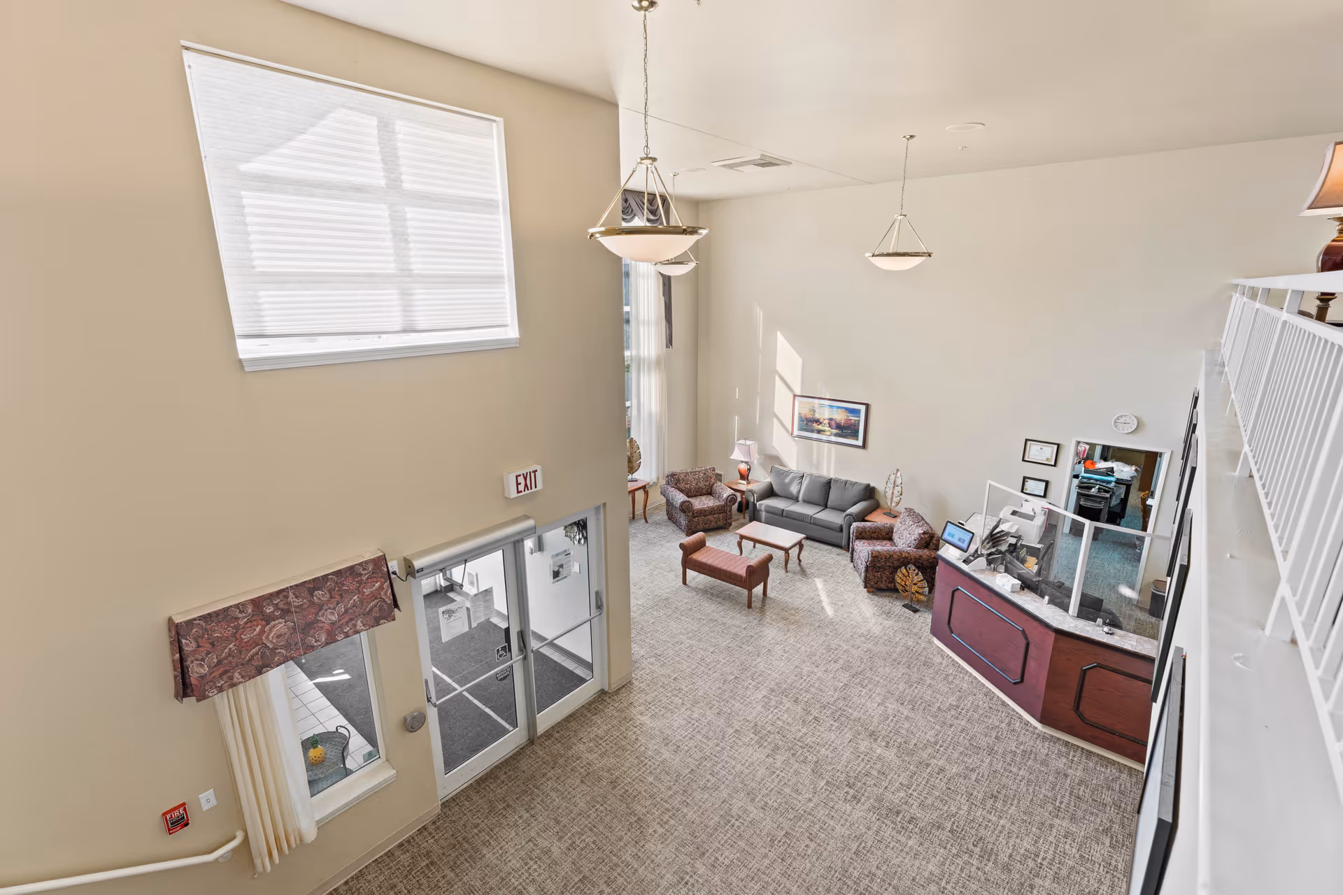 View from an upper level looking down into a bright assisted living facility lobby with a seating area including a gray sofa, two patterned armchairs, a coffee table, and a bench. There is a reception desk with a protective screen, a large window with blinds, and double glass doors marked with an exit sign. The walls are light-colored and the floor is carpeted.