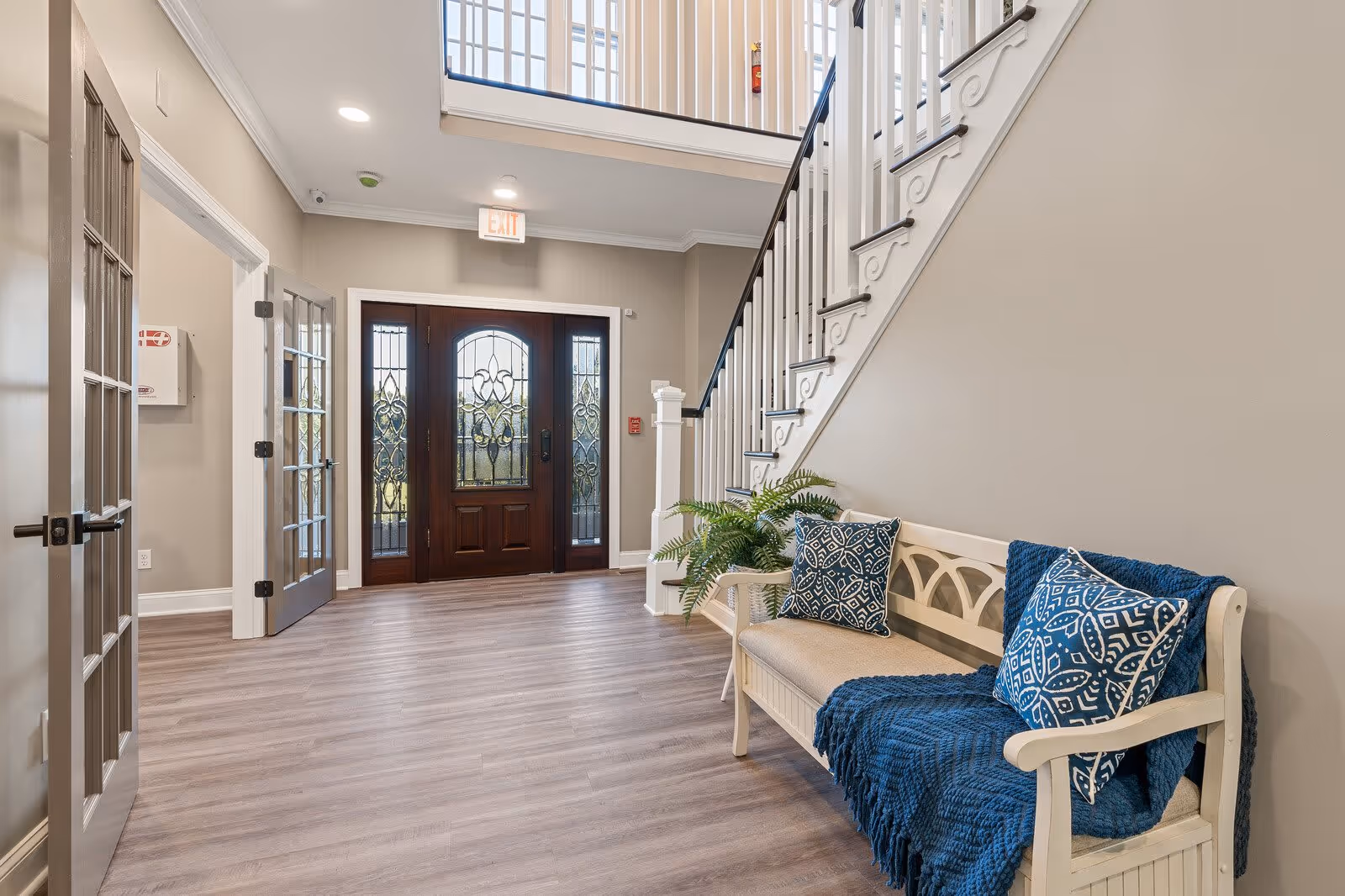 Interior view of a senior living facility entrance area with a wooden door featuring decorative glass panels, a staircase with white railings, a white bench with blue patterned pillows and a blue throw blanket, and a potted plant beside the bench. The floor is light wood, and there are double glass doors on the left side.