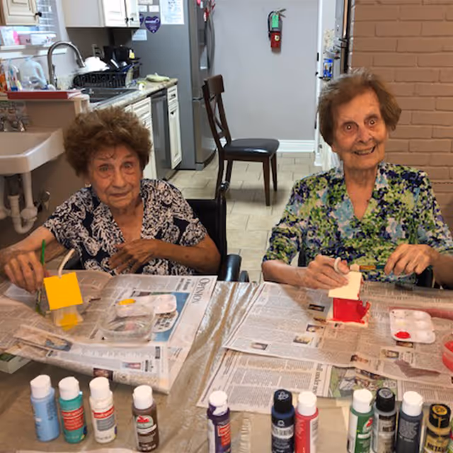 Two elderly women sitting at a table covered with newspapers, painting small wooden birdhouses. Various bottles of paint are lined up on the table in front of them. The setting appears to be a kitchen or dining area with a sink, cabinets, and a refrigerator in the background.