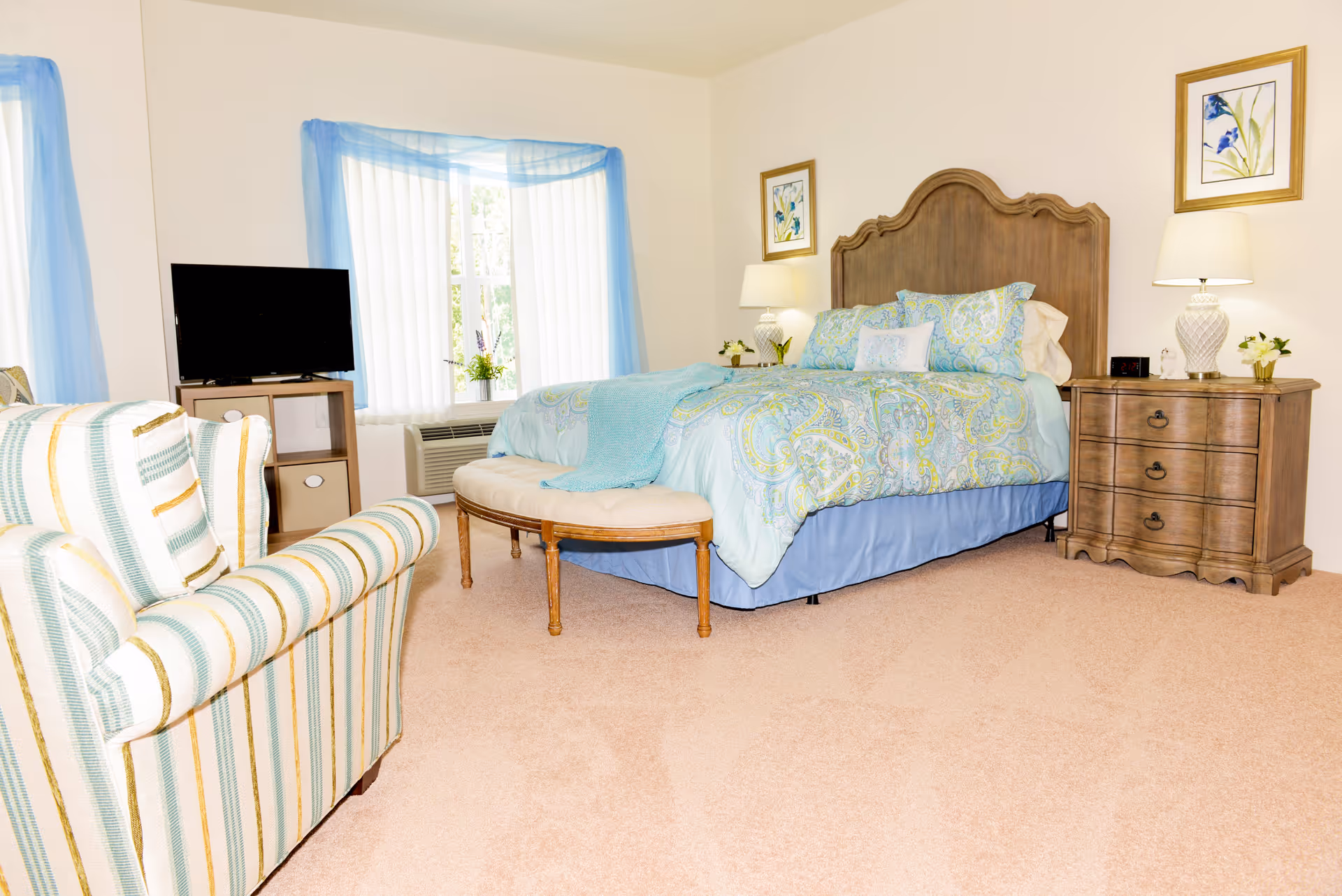 A bright bedroom with a large bed featuring a wooden headboard and blue patterned bedding. There is a cushioned bench at the foot of the bed, a wooden nightstand with a lamp and flowers, a striped armchair, a TV on a small stand, and windows with sheer blue curtains.