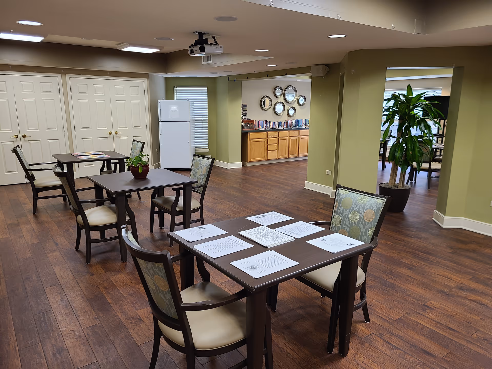A spacious room with wooden flooring featuring several tables and chairs arranged for activities. One table has papers and coloring sheets spread out. In the background, there is a white refrigerator, a set of double doors, a bookshelf with books, and decorative round mirrors on the wall. A large green plant is visible near an open doorway leading to another room.