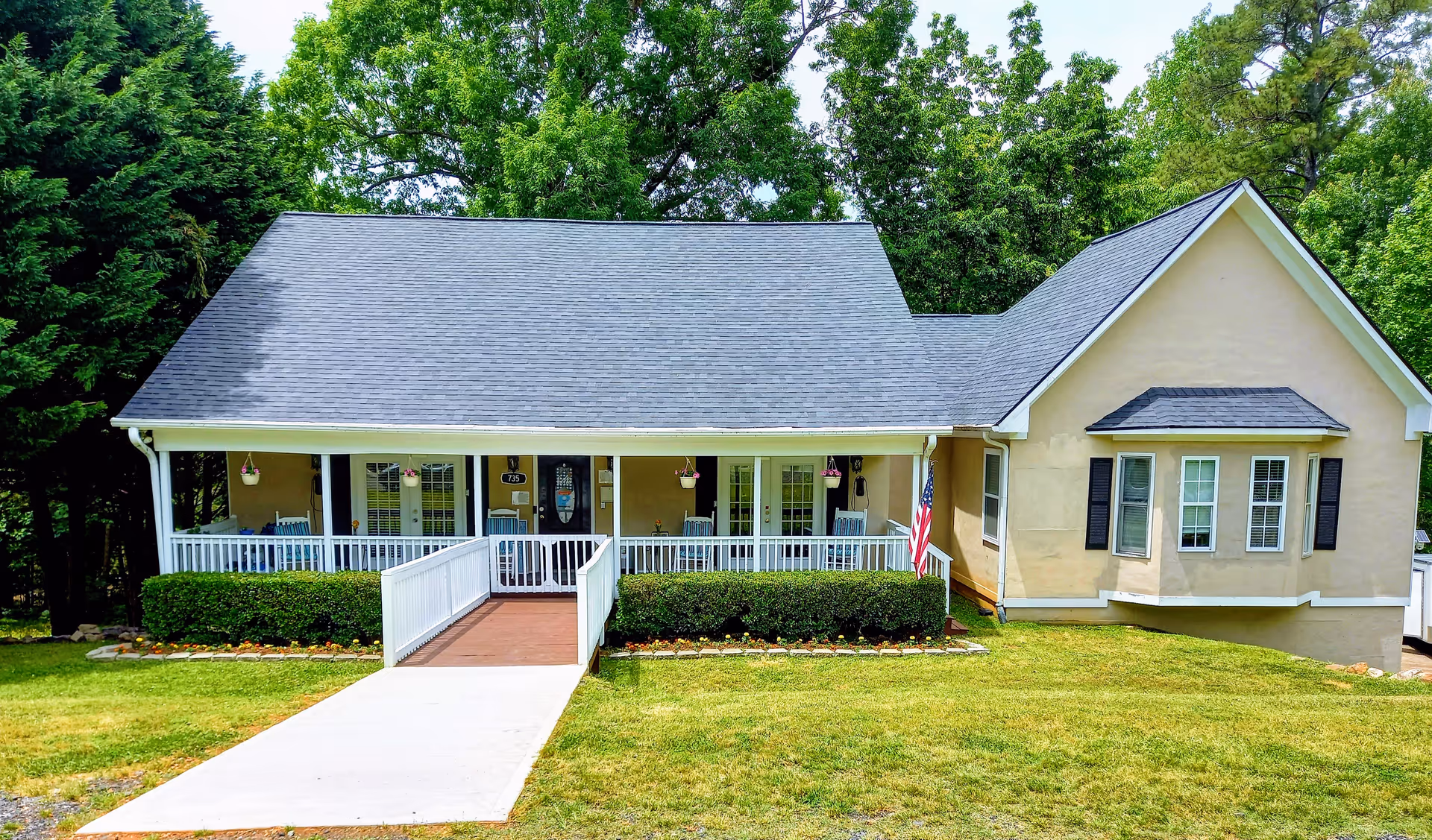 Front exterior view of a single-story building with a gray shingled roof, beige walls, and a white railing porch with a ramp leading to the entrance. There are hanging flower pots on the porch, an American flag on the right side, and green bushes along the front. The building is surrounded by green grass and trees in the background.