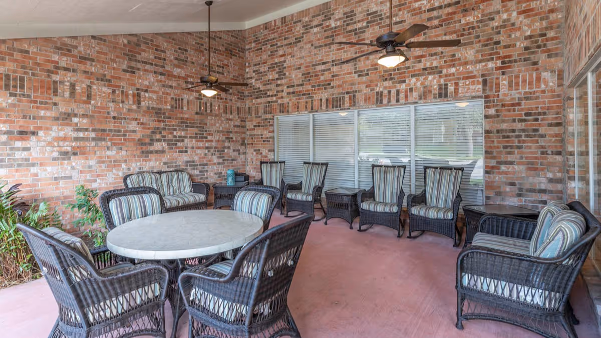 Covered outdoor patio area with brick walls, featuring a round table surrounded by four wicker chairs with cushions, additional wicker armchairs with striped cushions, ceiling fans with lights, and large windows with blinds.