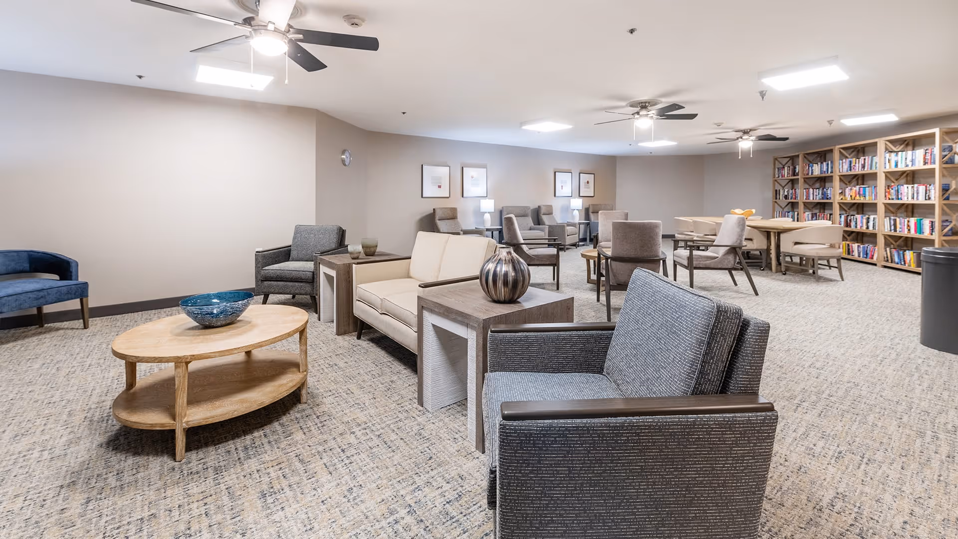 A spacious and well-lit common area with multiple seating options including armchairs and sofas arranged around wooden coffee tables. The room features ceiling fans, framed artwork on the walls, and a large bookshelf filled with books along one side. The carpeted floor and neutral wall colors create a cozy and inviting atmosphere.