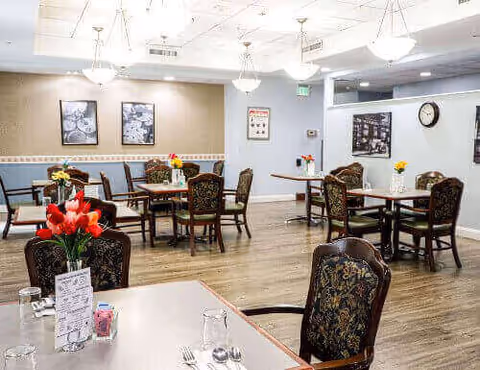 A bright dining room in a senior living facility with several tables and chairs arranged neatly. Each table has a small vase with colorful flowers. The room has wooden flooring, framed pictures on the walls, and ceiling lights providing ample illumination.