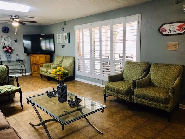 A cozy living room with a tiled floor, two green patterned armchairs, a glass coffee table with a vase of yellow flowers, a flat-screen TV on a wooden stand, a ceiling fan with lights, and a large window with white plantation shutters.