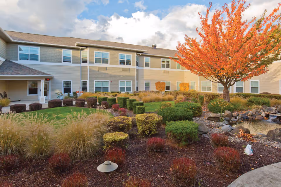 Landscaped courtyard and pond in front of a two-story senior living building with an orange-leaved tree in autumn.