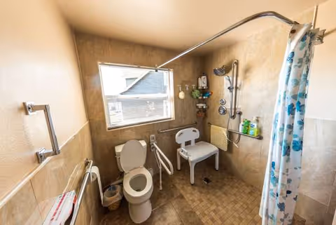 Accessible bathroom with toilet, shower chair, grab bars, and patterned curtain.