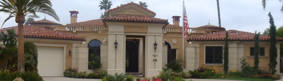 Front exterior view of a single-story building with beige walls, red-tiled roof, an American flag on a pole, and surrounding palm trees and greenery.