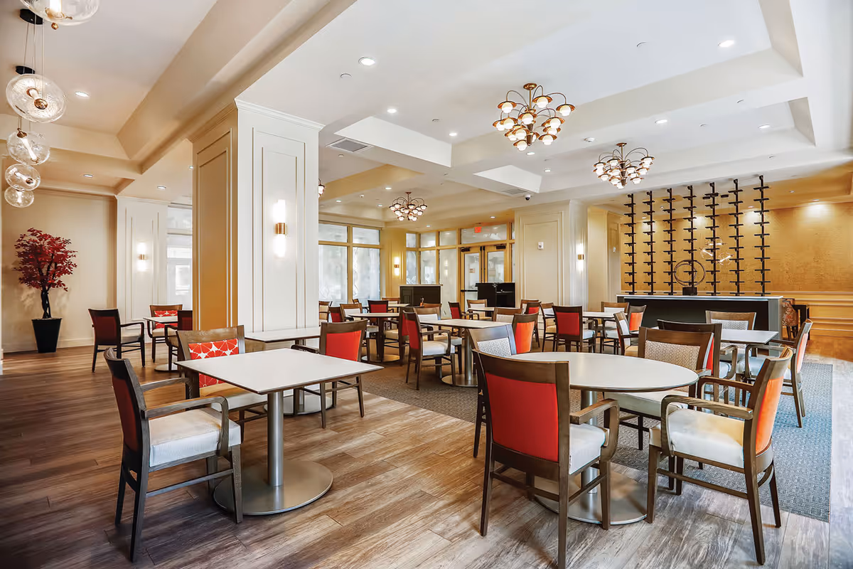 A spacious and well-lit dining room with multiple tables and chairs arranged neatly. The chairs have wooden frames with red and beige cushions. The room features modern chandeliers, large windows allowing natural light, and a decorative wine rack on one wall. There is a potted plant with red leaves in the corner, and the flooring is a combination of wood and carpet.