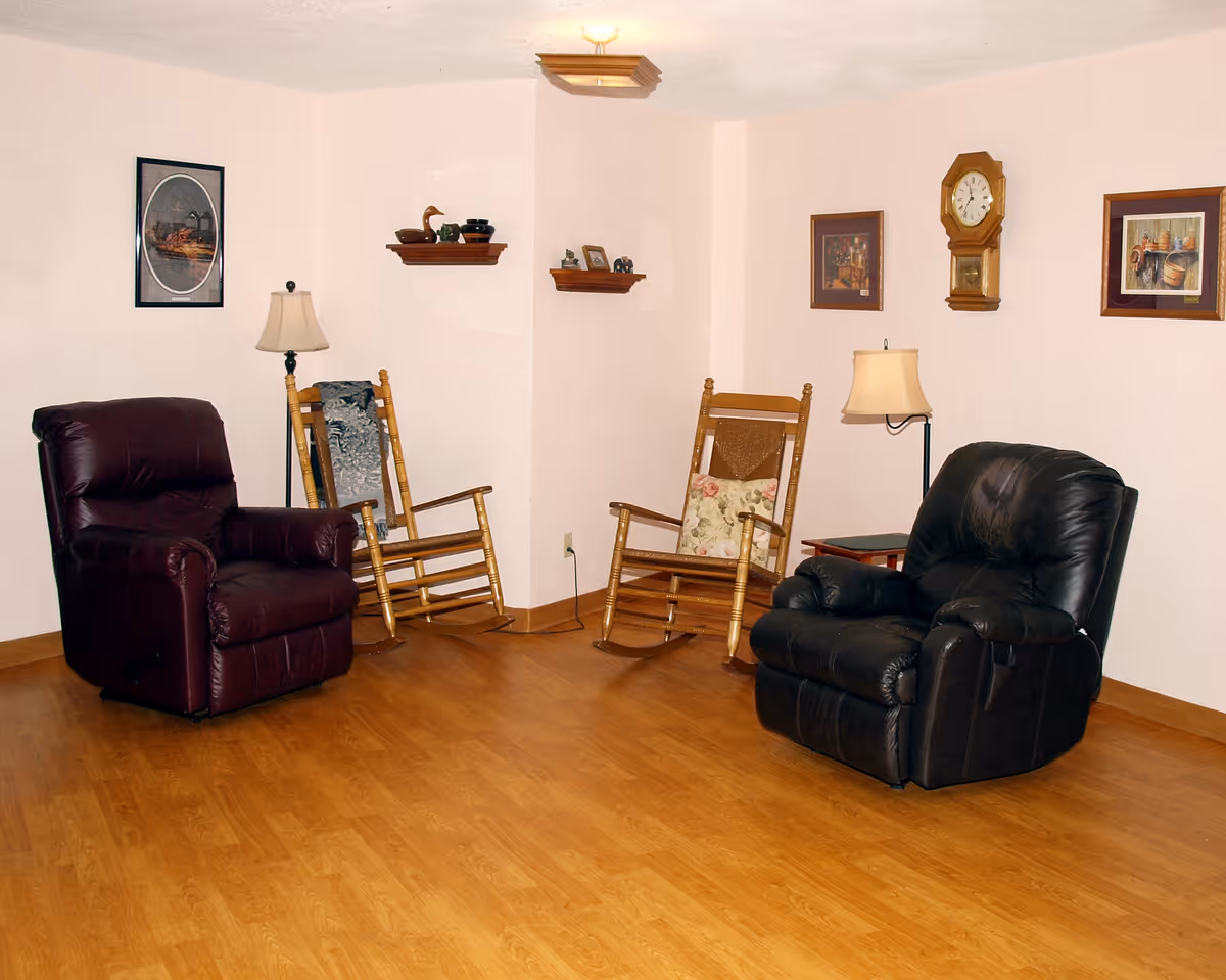 A cozy sitting area with two leather recliners and two wooden rocking chairs arranged on a wooden floor. The walls are light-colored and decorated with framed pictures, a wall clock, and small shelves holding decorative items. Two floor lamps provide lighting.