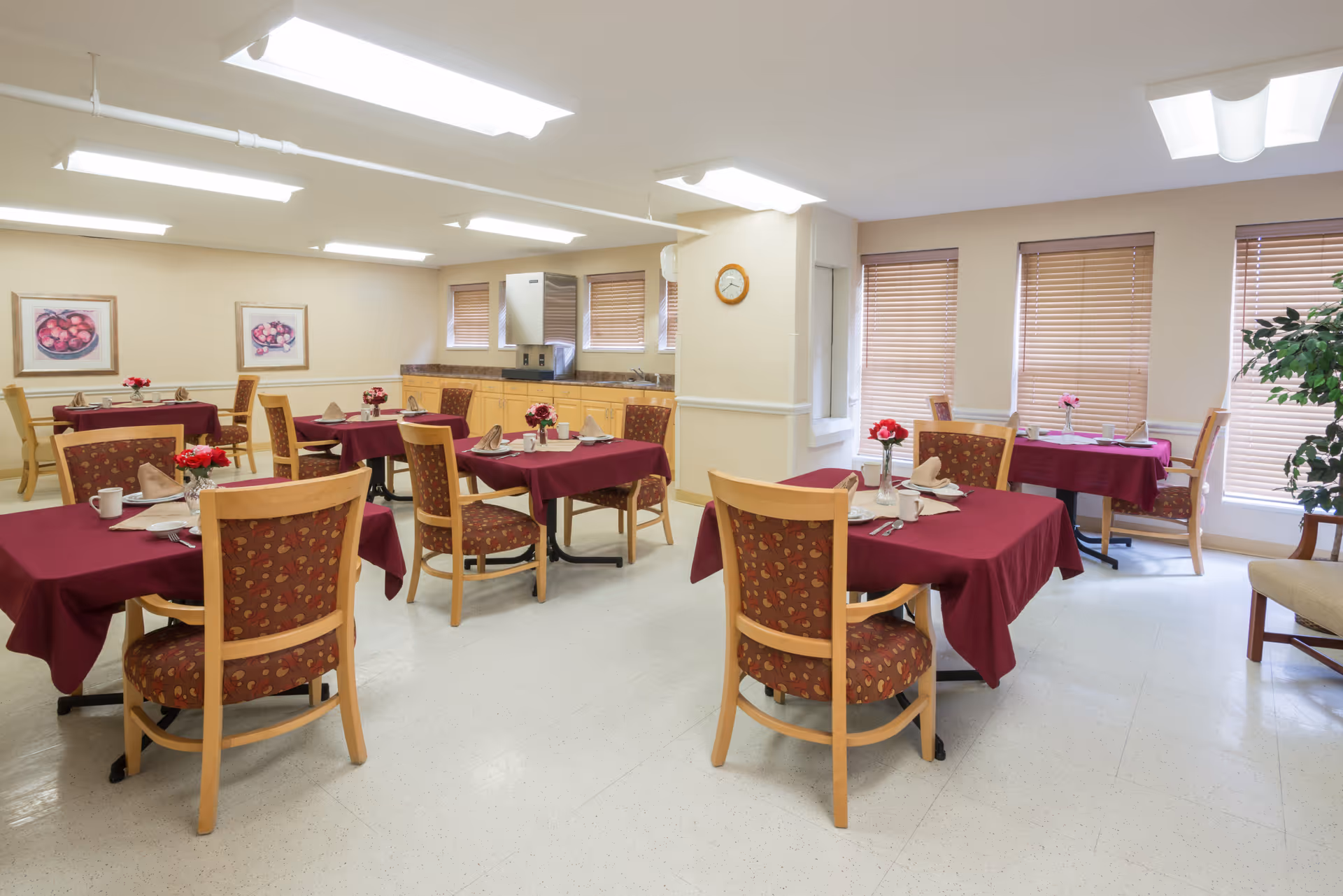 A bright dining room with several tables covered in burgundy tablecloths, each set with napkins, cups, and small flower arrangements. The room has wooden chairs with patterned upholstery, large windows with blinds, and light-colored walls adorned with framed artwork. There is a kitchenette area with cabinets, a microwave, and a clock on the wall.