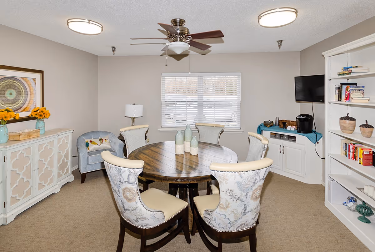 A cozy interior room with a round wooden table surrounded by six upholstered chairs with floral patterns. The room features a ceiling fan with lights, a window with blinds, a white cabinet with a coffee maker and cups, a bookshelf with books and decorative items, a blue armchair with a bird-patterned pillow, and a white sideboard with two vases of sunflowers and a framed artwork above it.