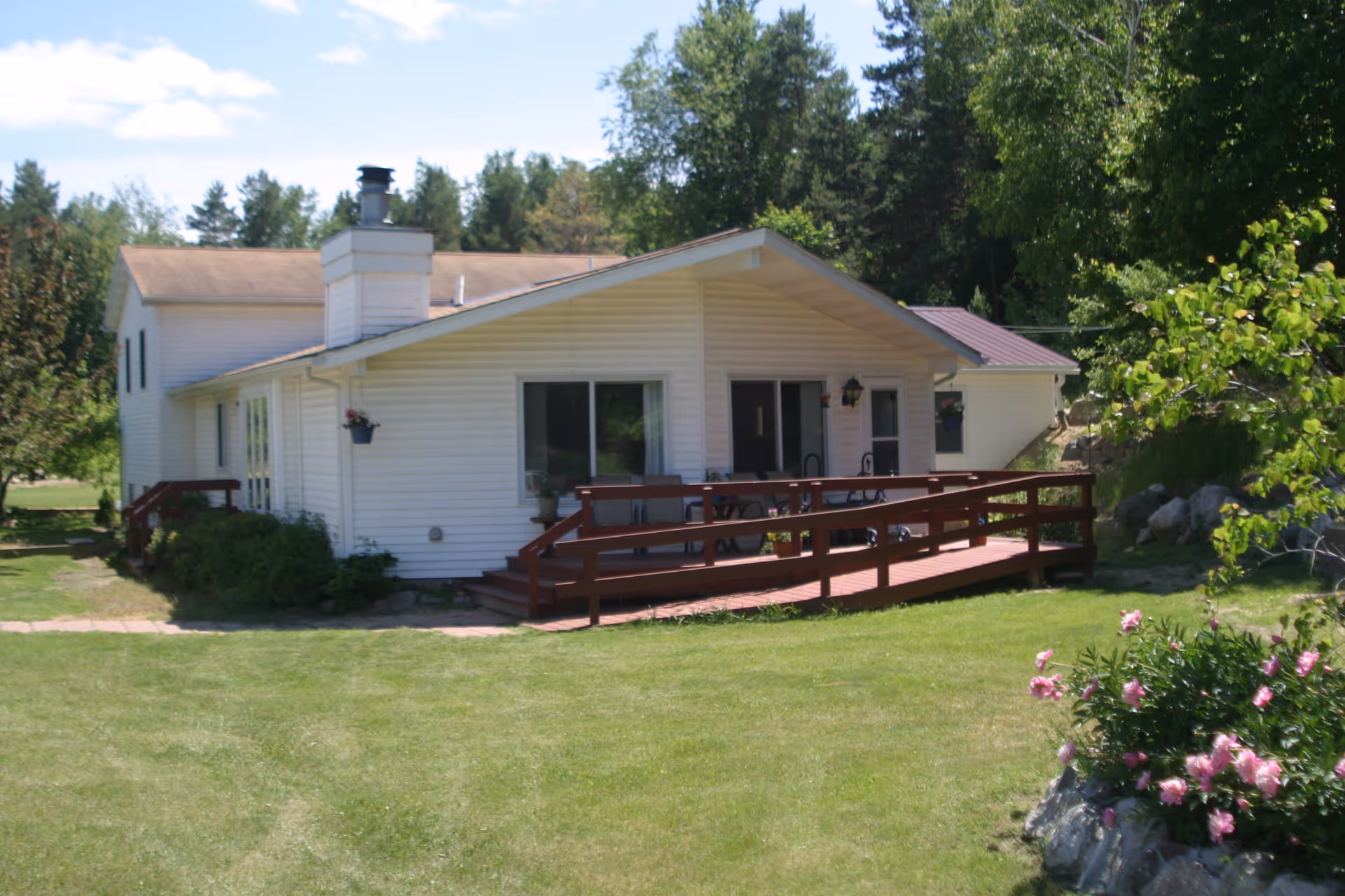 Exterior view of a single-story white building with a brown roof and a wooden ramp leading to a porch area. The building is surrounded by green grass, trees, and flowering plants under a partly cloudy sky.