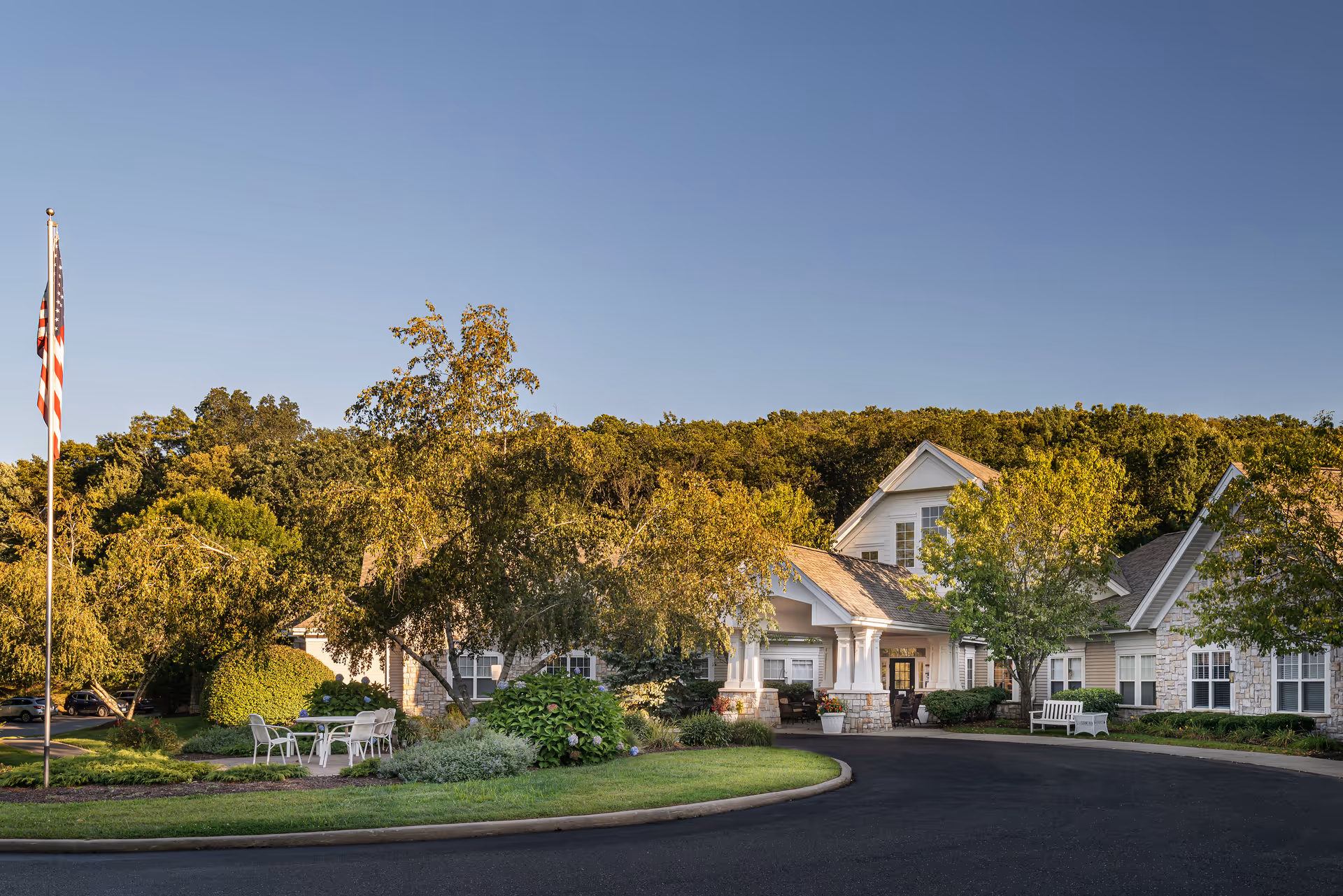 Front entrance of a memory care building with a circular driveway, flagpole, outdoor seating, and landscaped trees and shrubs.