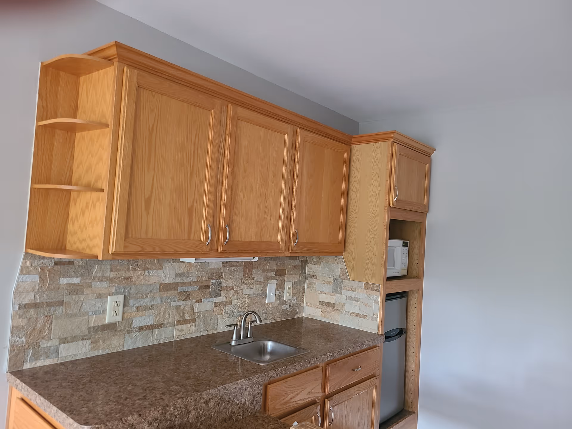A kitchen area with wooden cabinets, a small stainless steel sink with a faucet, a brown countertop, and a stone tile backsplash. There is a microwave and a mini refrigerator built into the cabinetry on the right side. The walls are painted light gray.