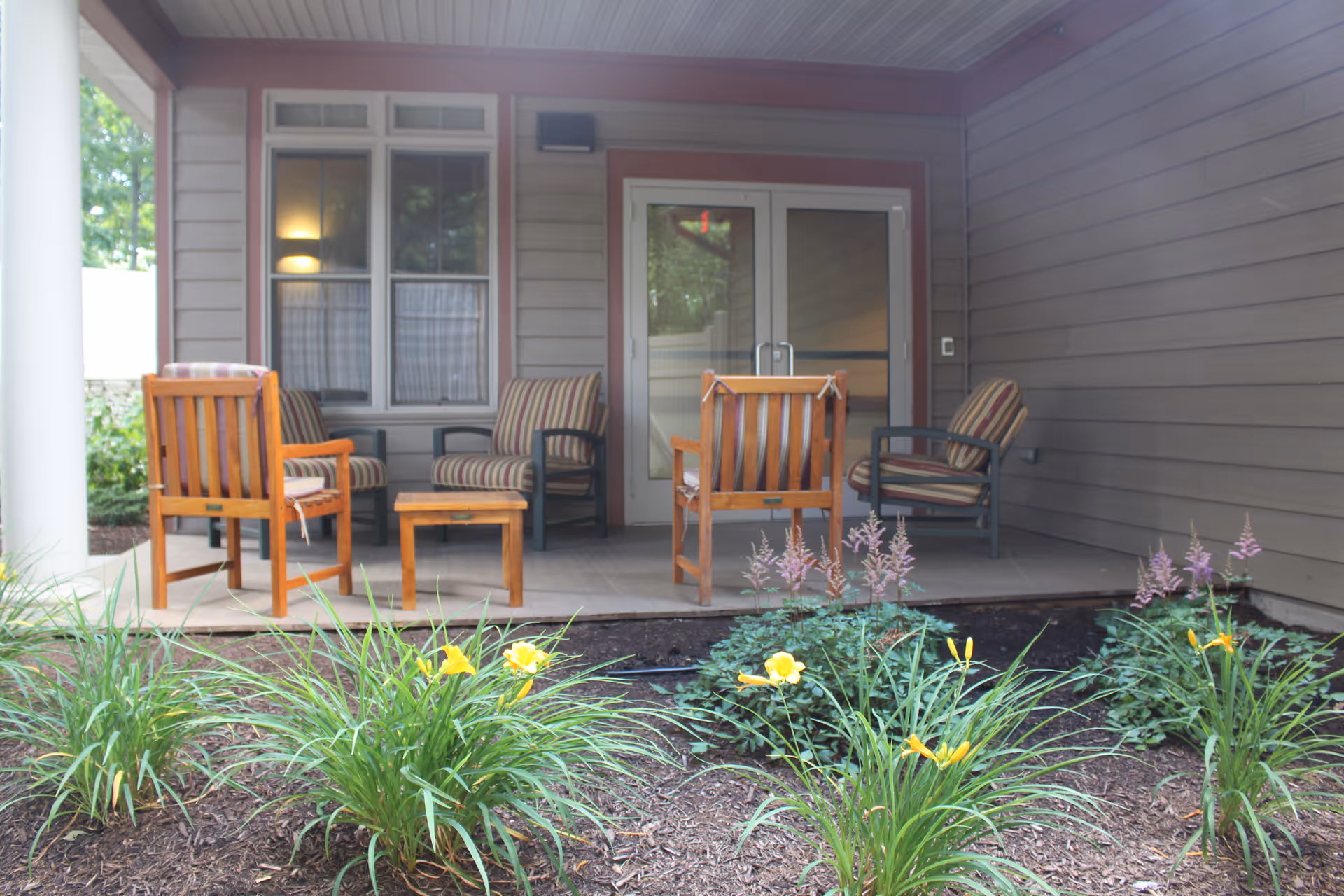 Covered porch with wooden chairs and a small table in front of sliding glass doors and a flower bed with yellow flowers.