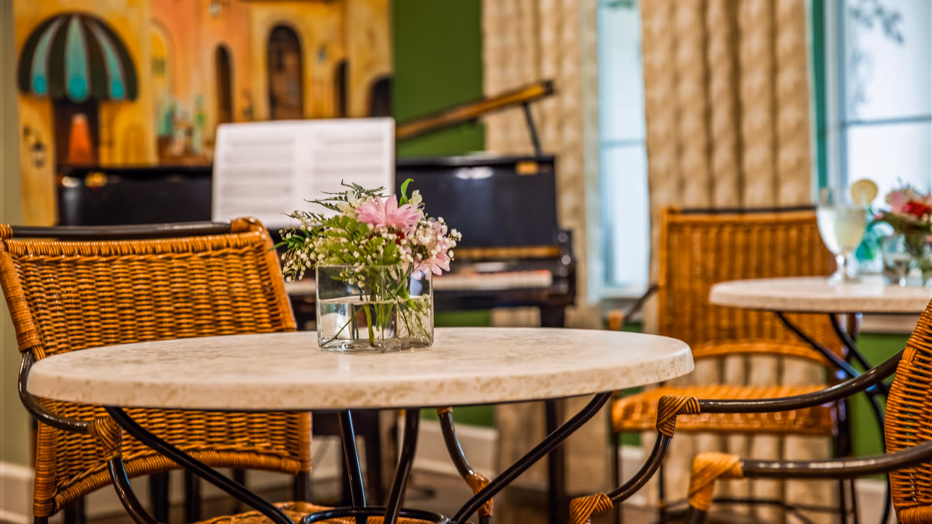 A cozy interior dining area with round marble tables and wicker chairs. Each table has a small glass vase with fresh flowers. In the background, there is a black piano with sheet music on the stand, and a window with beige curtains letting in natural light.