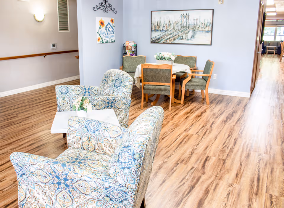 A cozy seating area in a senior living facility with two patterned armchairs around a small white table with a flower arrangement. In the background, there is a round table with four green cushioned chairs and a floral centerpiece. The walls are light blue with a framed picture of a bridge and a decorative wall sign that reads 'Our House Senior Living'. The floor is wood-patterned laminate.