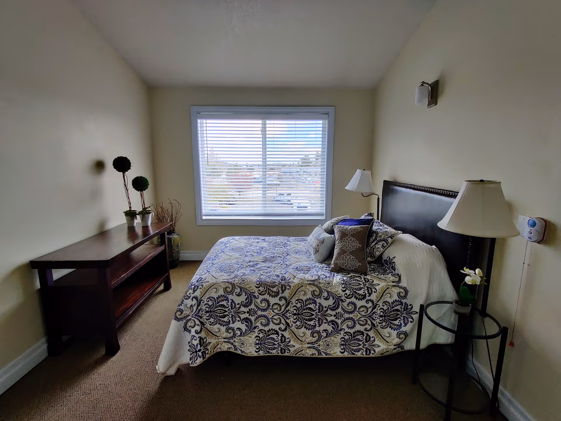 A bedroom with a bed covered in a white and blue patterned bedspread, several decorative pillows, a dark wooden headboard, a round glass side table with a lamp and a small plant, a wooden console table with decorative plants, and a large window with blinds letting in natural light.