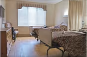 Shared nursing facility room with two hospital beds, a dresser and TV, and a large window with blinds.