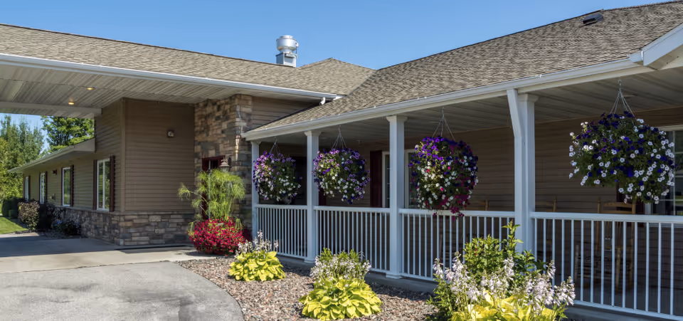 Exterior view of a senior living facility building with a covered porch featuring hanging flower baskets and landscaped garden beds with various plants and flowers under a clear blue sky.