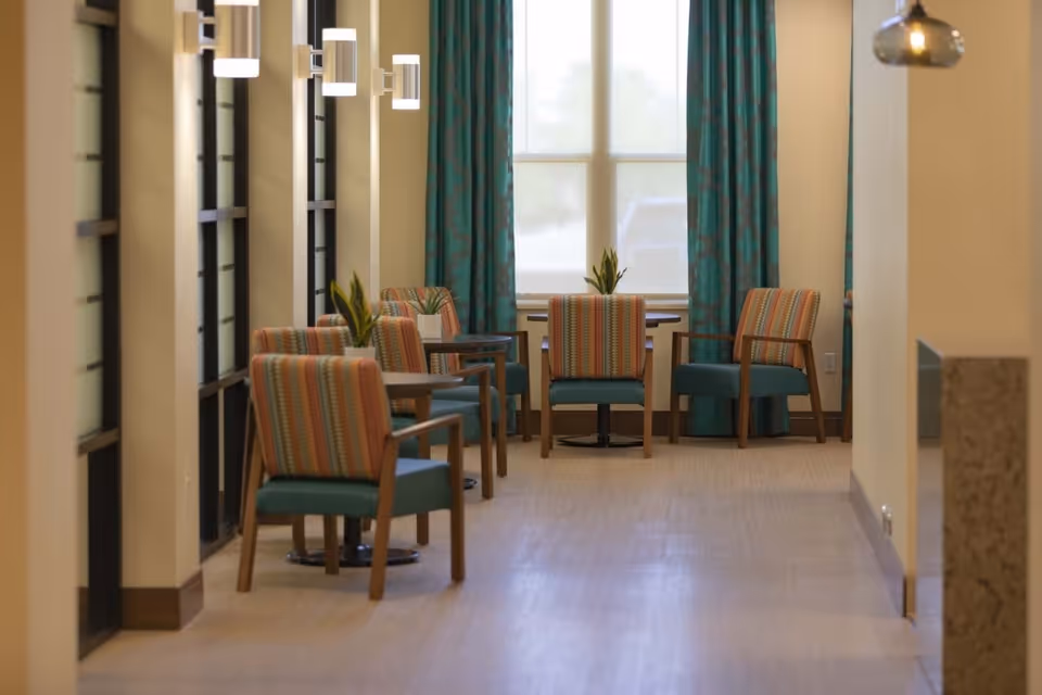 A hallway seating area with several wooden chairs featuring colorful striped cushions and teal seats, small tables with potted plants, teal curtains on the window at the end, and modern wall-mounted lights along the left side.