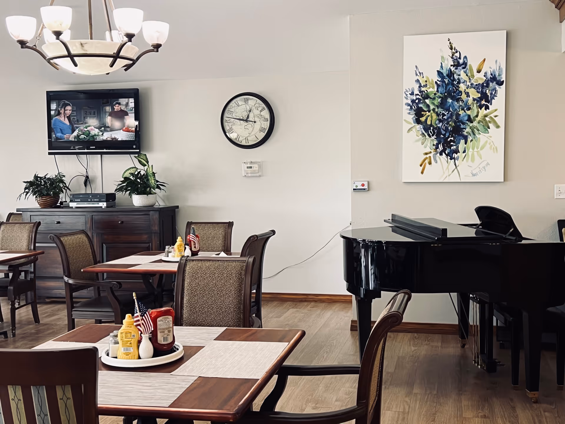 Dining room with tables and chairs, a grand piano, a wall-mounted TV, clock, and floral artwork.
