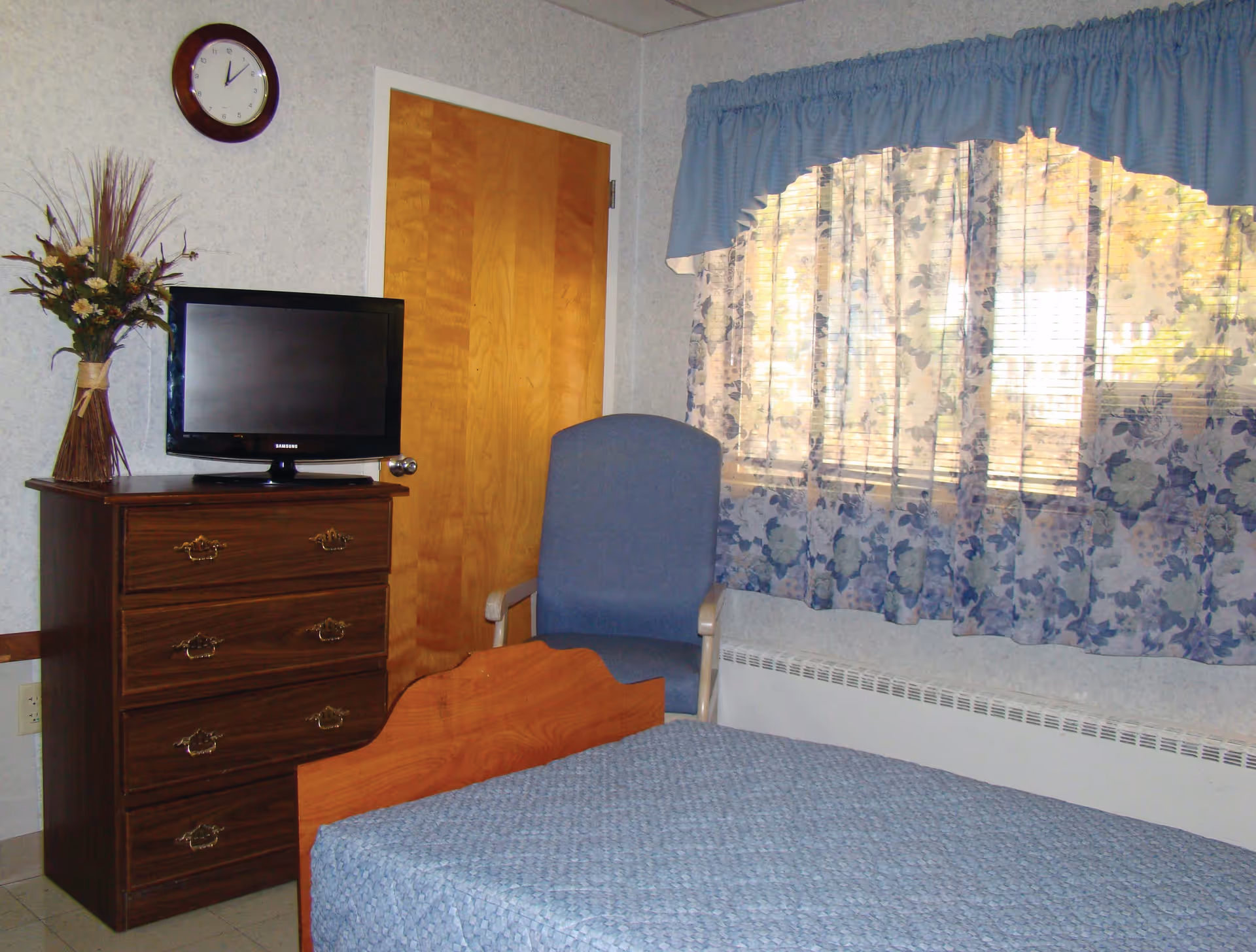 A simple bedroom with a wooden bed covered in a blue quilt, a wooden dresser with a small flat-screen TV on top, a blue upholstered chair, a window with floral curtains and a blue valance, a wooden door, and a wall clock showing 12:05.