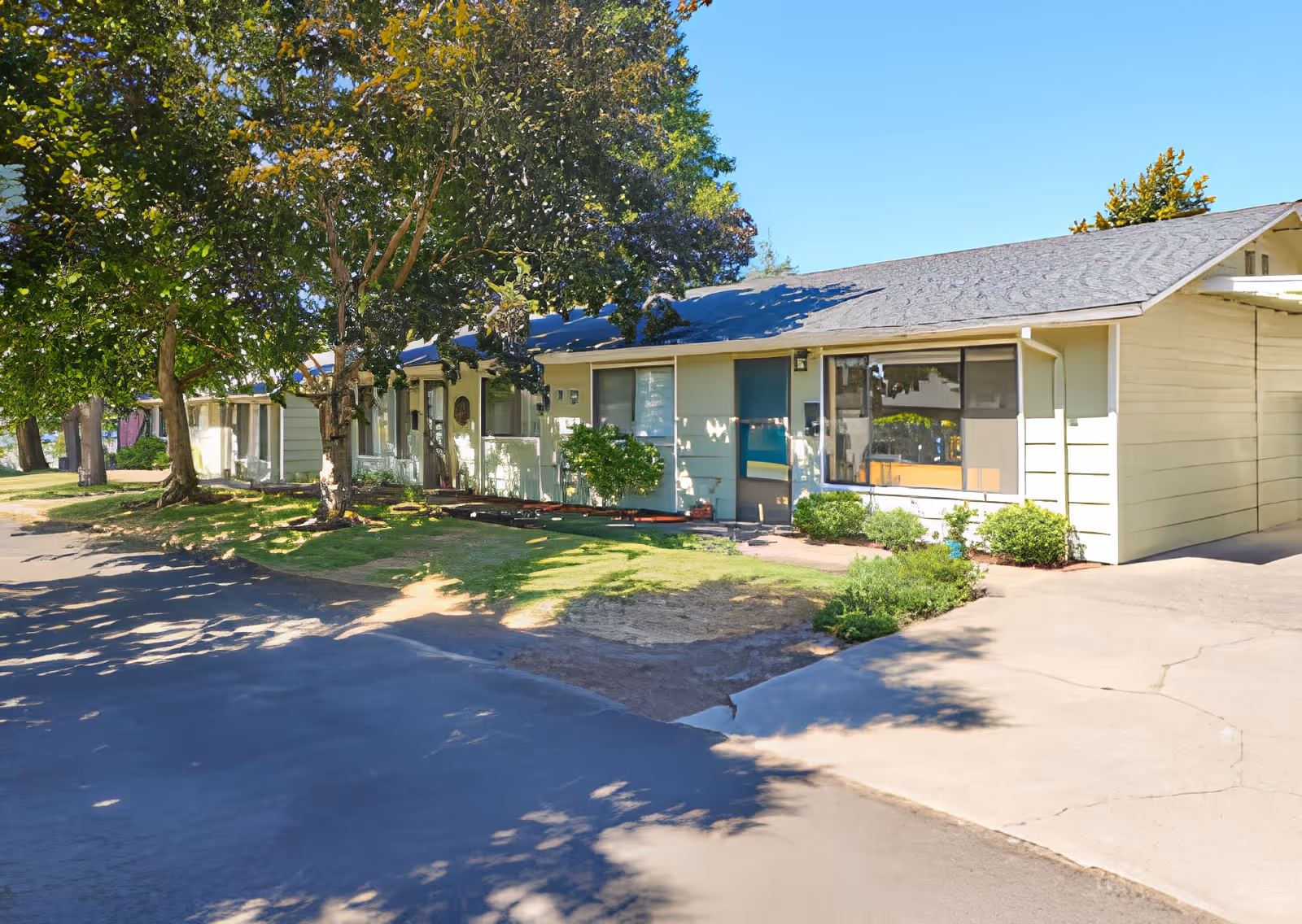 Single-story light-colored residential building with several ground-level units, trees, front lawn and a driveway.