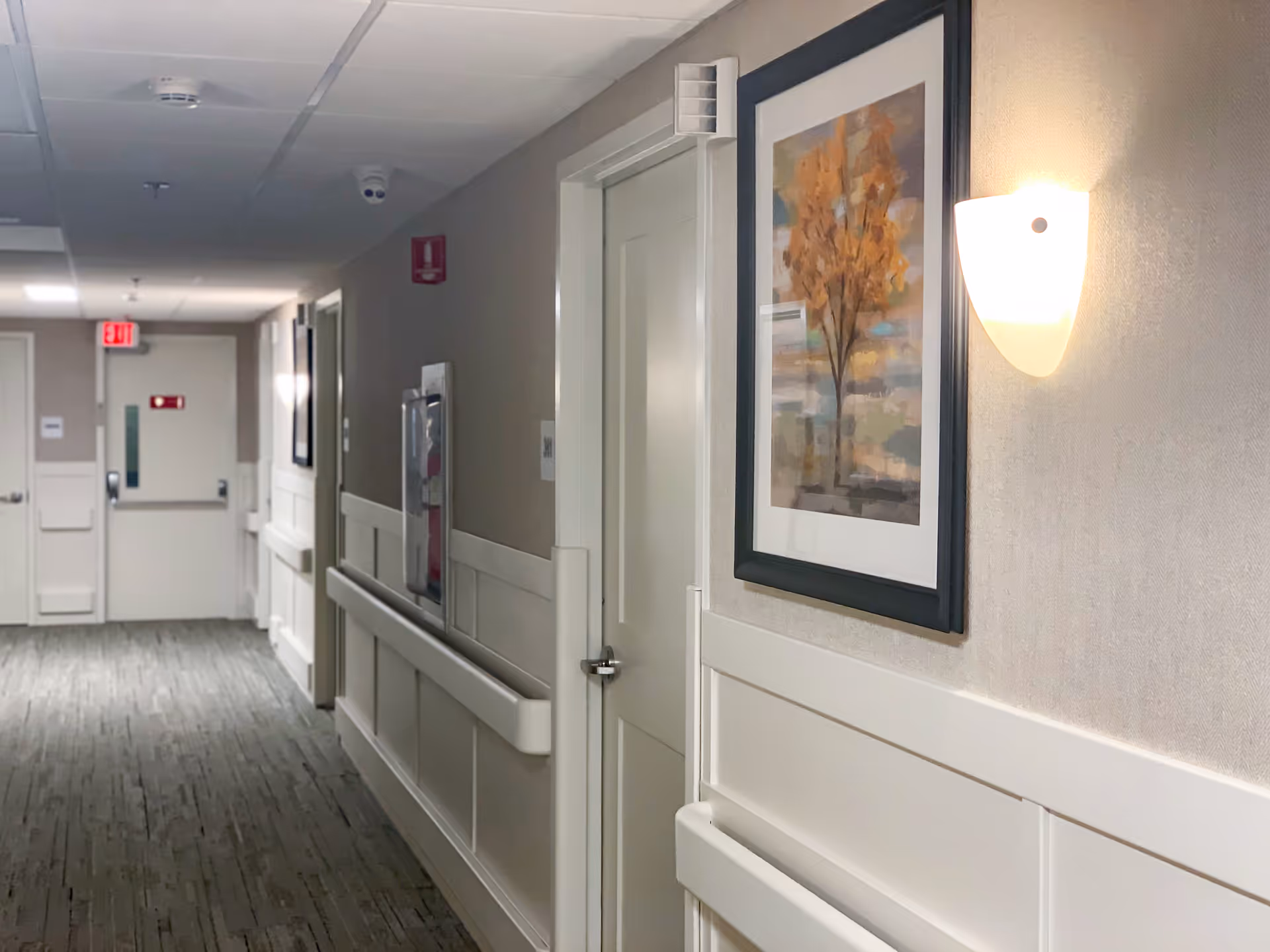 A well-lit hallway in a skilled nursing and rehabilitation center with beige walls, white doors, handrails along the walls, a framed painting of a tree with autumn leaves, and a wall-mounted light fixture. The hallway has a carpeted floor and an exit sign visible at the far end.