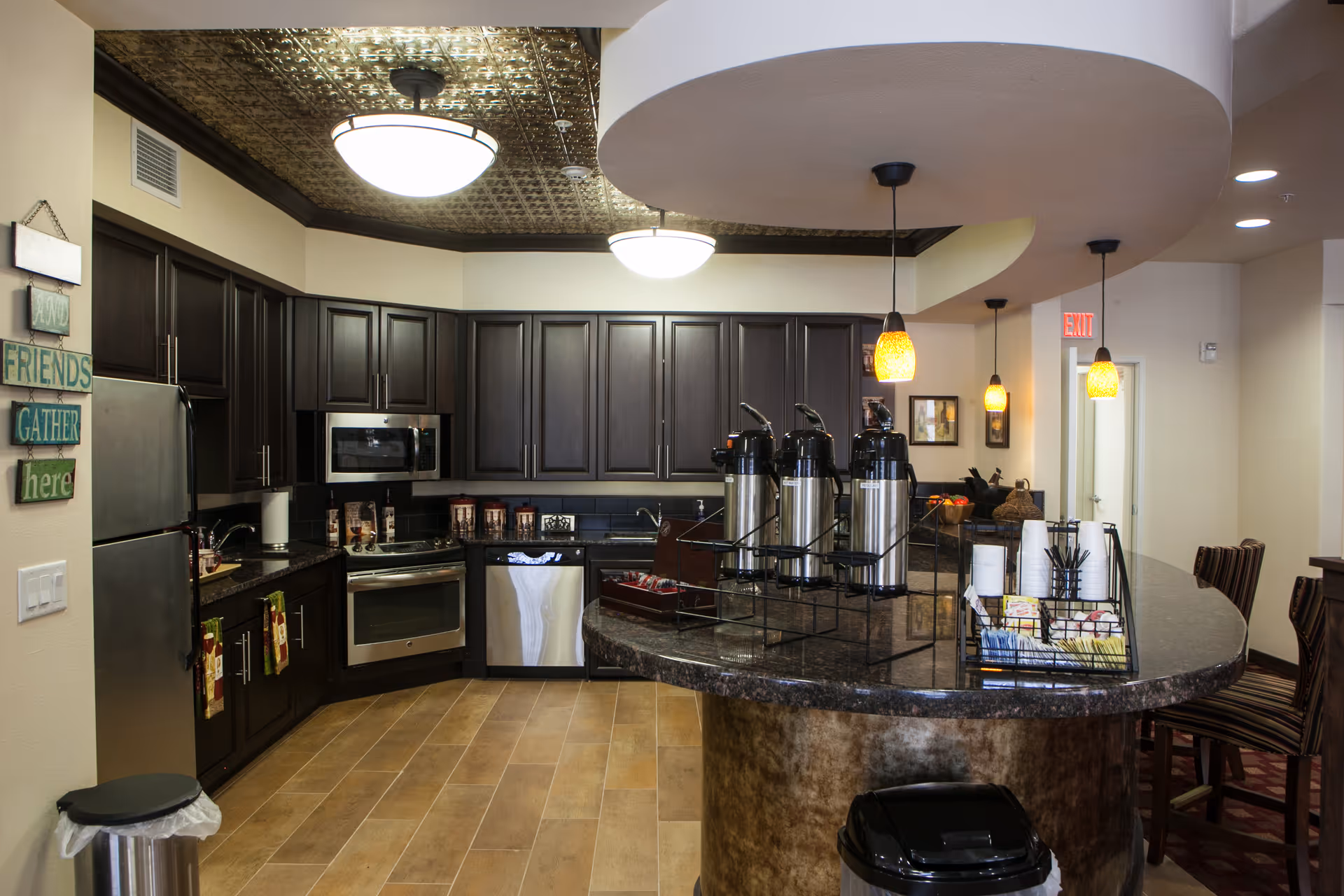 A modern kitchen area with dark wood cabinets, stainless steel appliances including a refrigerator, microwave, oven, and dishwasher. The kitchen has a curved granite countertop island with three large coffee dispensers and a basket holding cups and condiments. There are pendant lights hanging above the island and a decorative ceiling light. A sign on the wall reads 'AND FRIENDS GATHER here'.