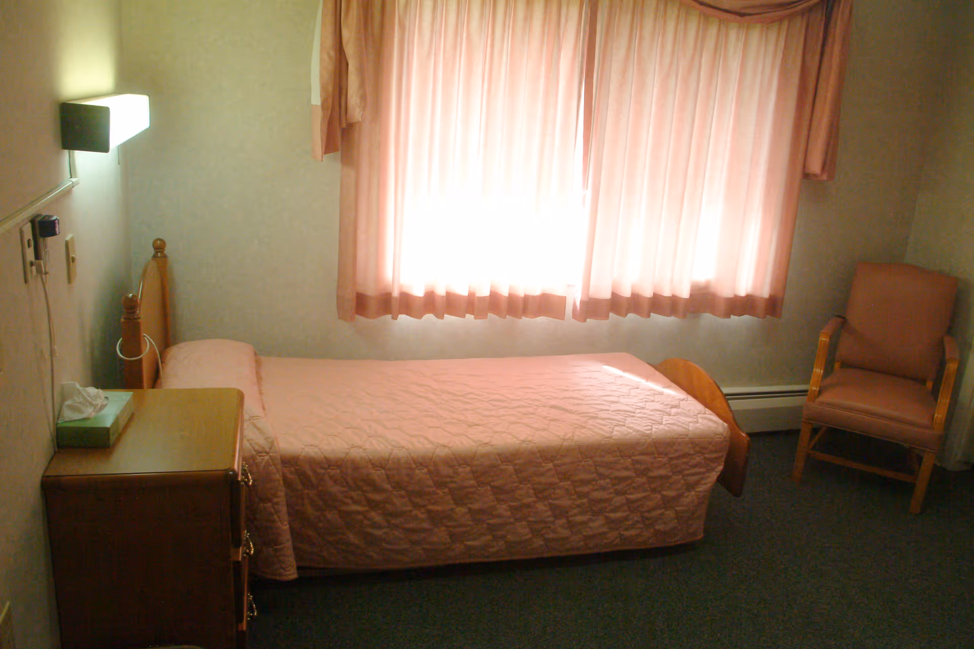 A small bedroom with a single bed covered in a pink quilted bedspread, a wooden nightstand with a box of tissues, a wall-mounted light above the bed, a window with pink curtains letting in light, and a pink upholstered wooden chair in the corner.