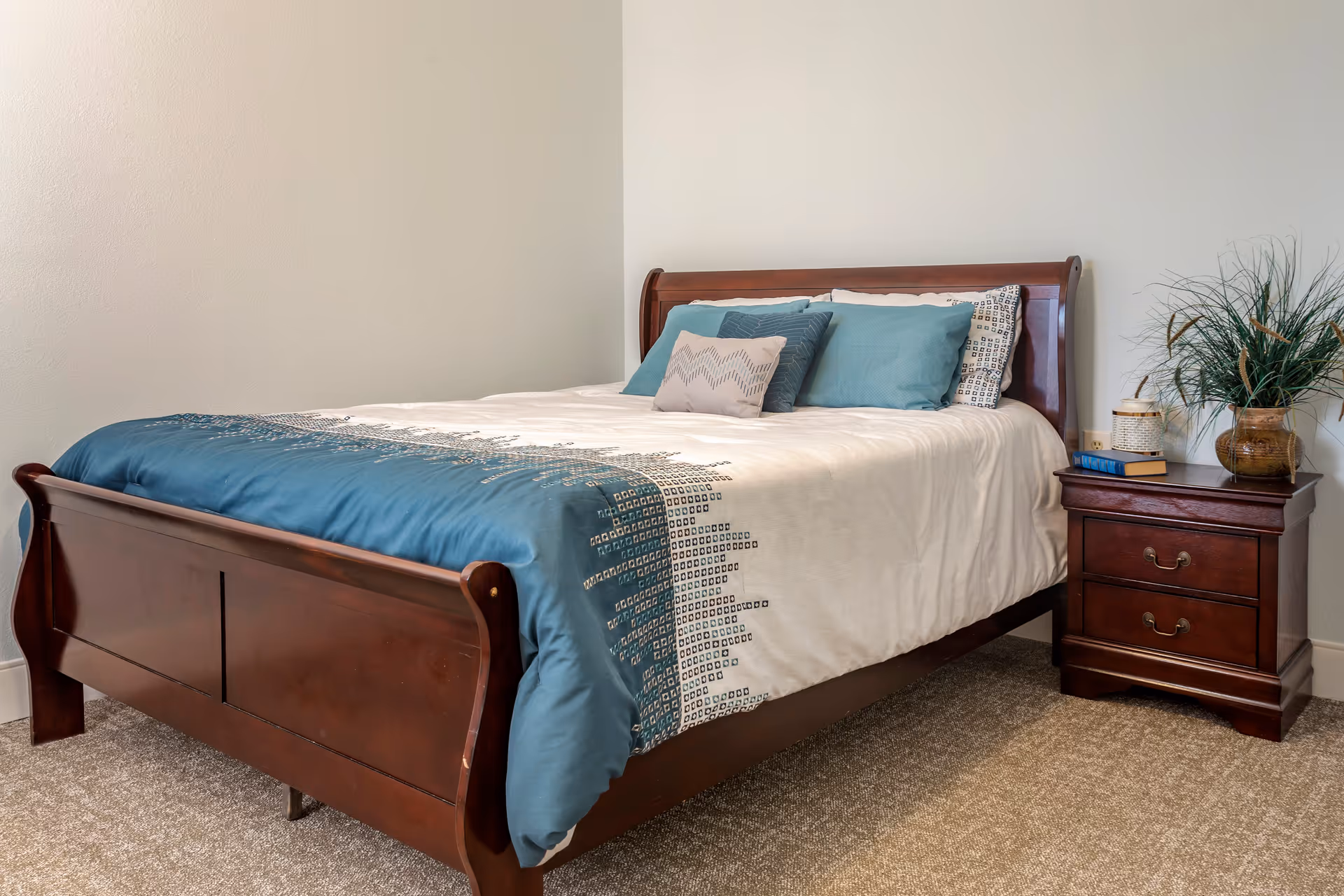 A neatly made bed with teal and white bedding and a wooden nightstand with a plant in a bright bedroom.