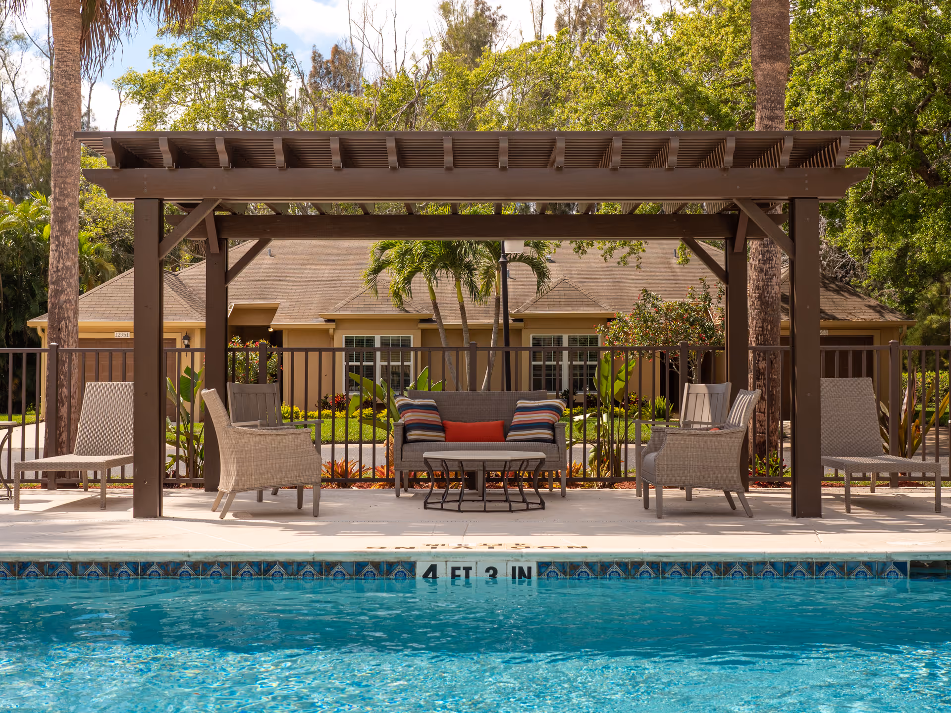 Pergola-covered seating area with chairs and a sofa beside a swimming pool in front of a residential building.