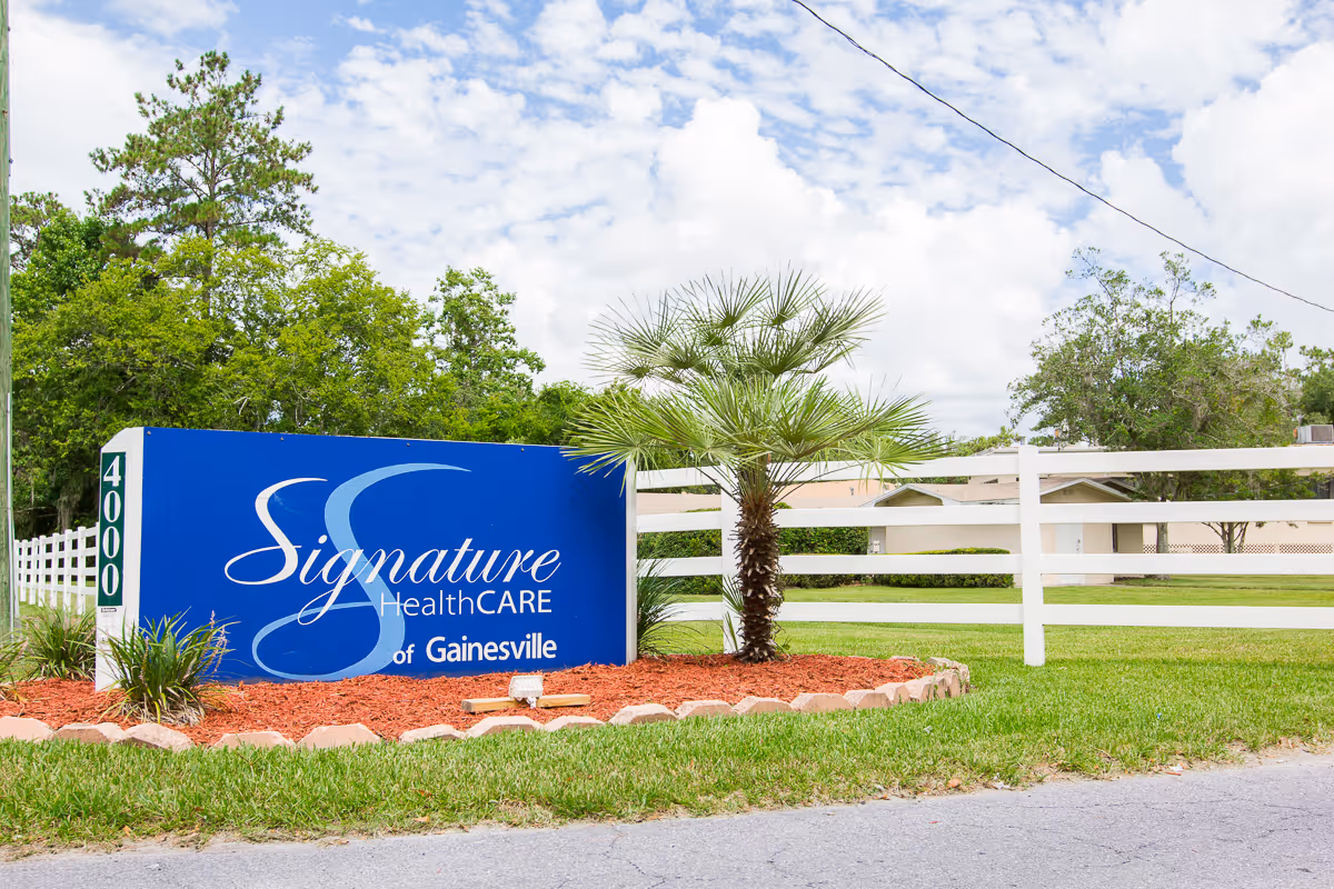 Blue 'Signature HealthCARE of Gainesville' sign in front of a white fence and landscaped lawn with a palm tree.