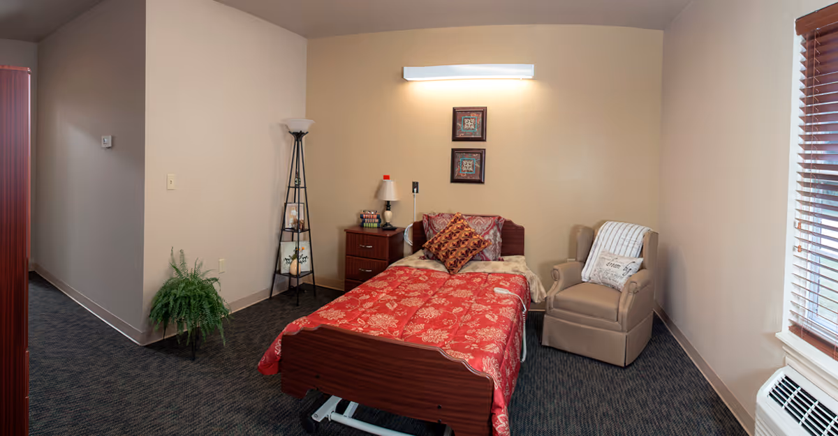 A cozy bedroom in a memory care facility featuring a single bed with a red floral comforter and decorative pillows. Next to the bed is a wooden nightstand with a lamp and some items on it. A tall floor lamp with shelves stands in the corner, holding framed pictures and a small plant. There is a comfortable beige armchair with a throw blanket and pillow near a window with wooden blinds. The room has beige walls and carpeted flooring.