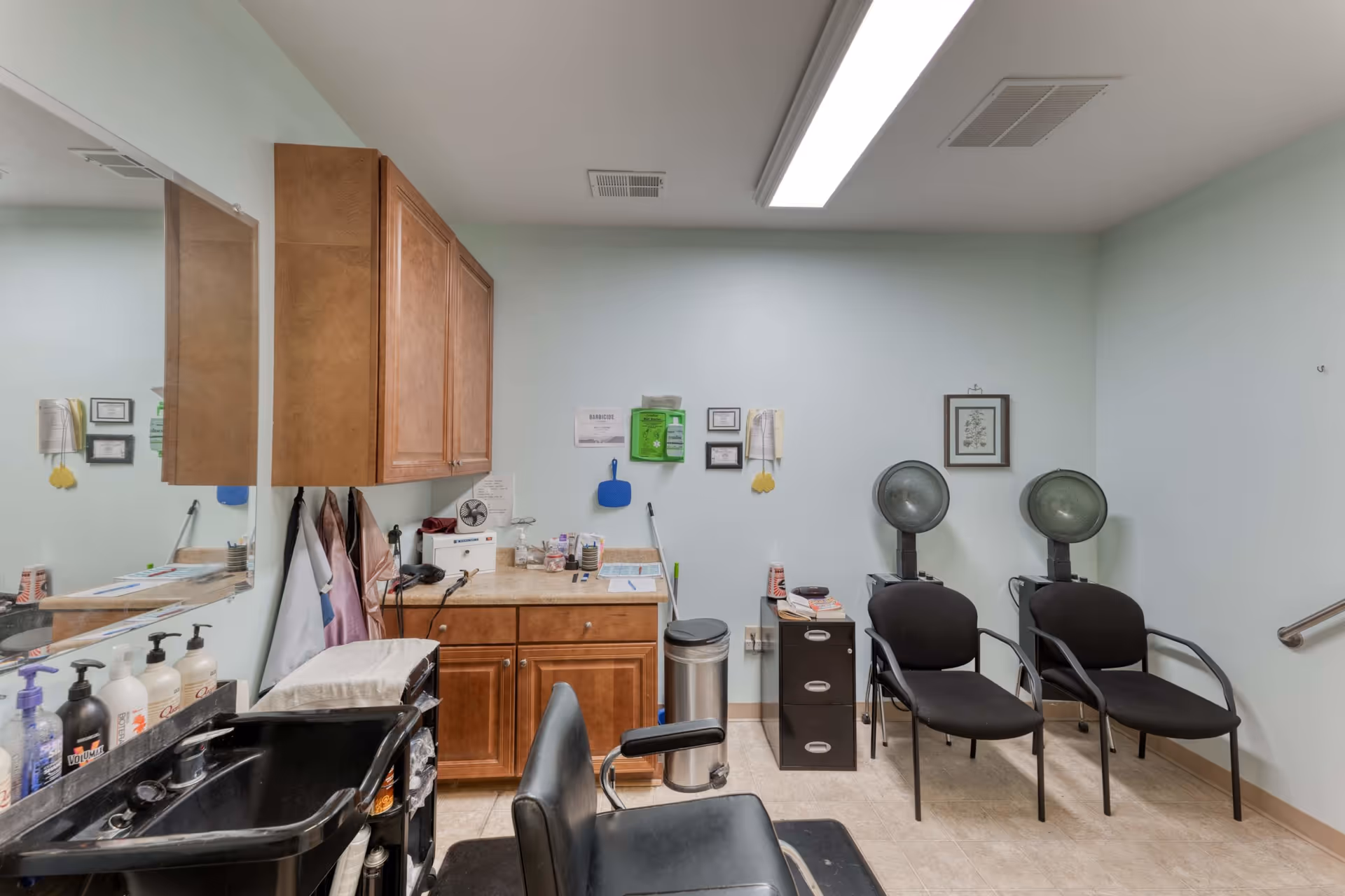 Interior view of a salon area in a senior living facility with a black salon chair in front of a black sink, wooden cabinets on the wall, a countertop with various supplies, two black chairs with hair dryers, and framed pictures on the light blue walls.