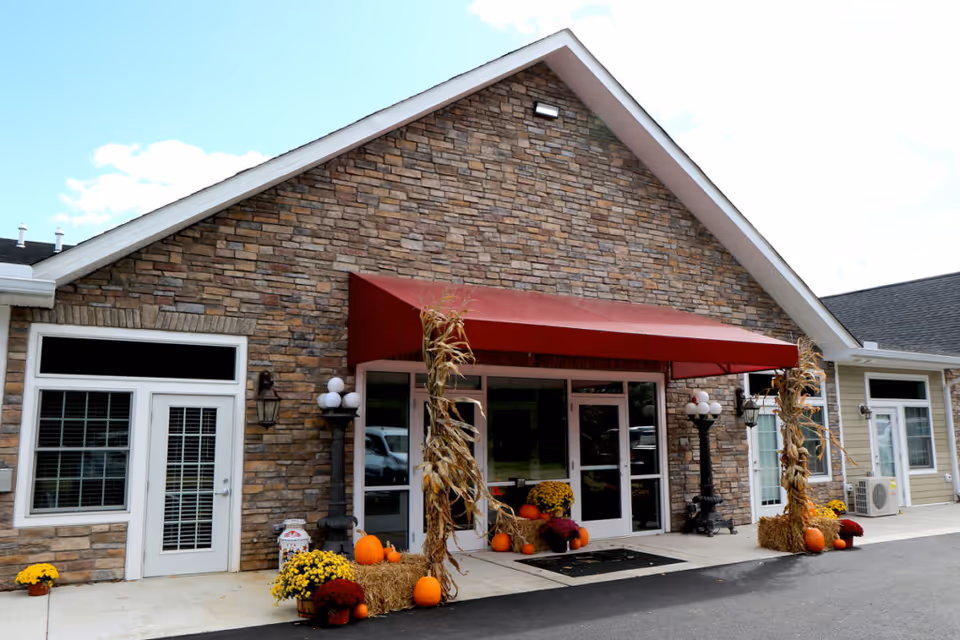 Exterior front entrance of a building with stone facade and a red awning above glass double doors. The entrance is decorated with autumn-themed items including pumpkins, hay bales, corn stalks, and potted flowers. There are white-framed windows and doors on either side of the entrance, and black lamp posts with globe lights near the doorway.