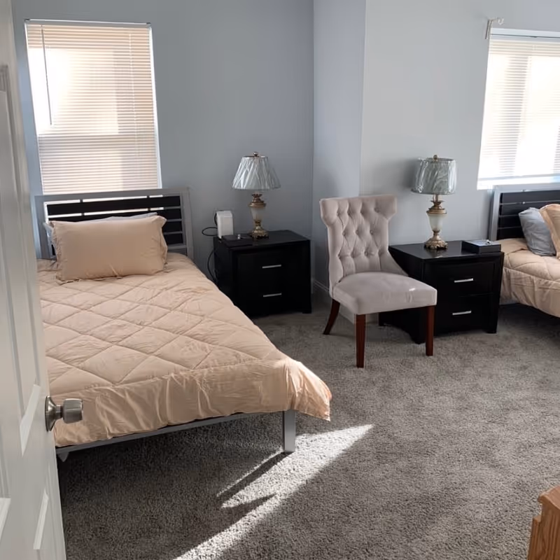 Two twin beds with nightstands, a tufted chair, and lamps in a light blue bedroom.