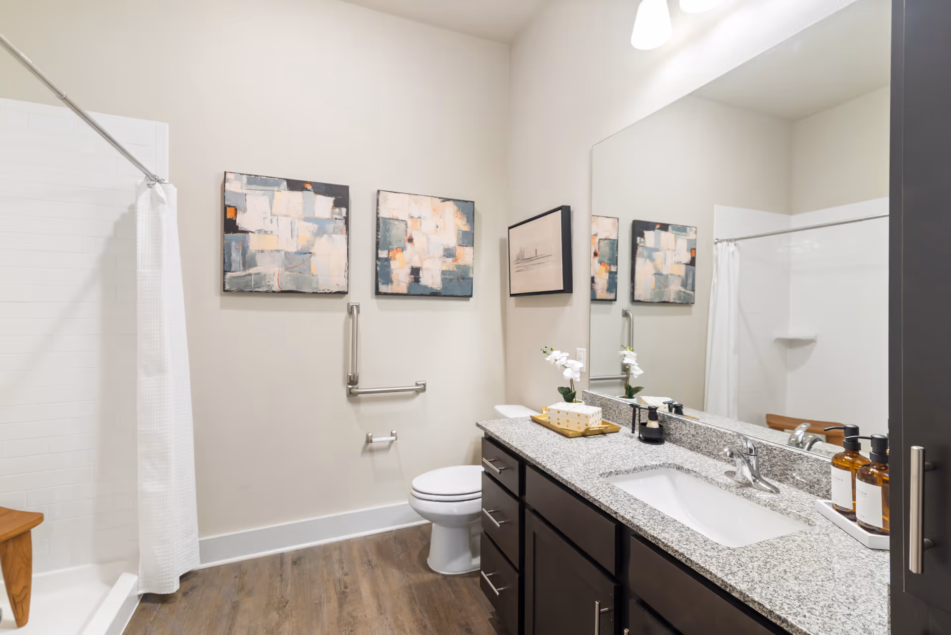 Well-lit accessible bathroom with a shower and curtain, toilet, granite countertop sink with dark cabinets, large mirror, wall art, and grab bars.