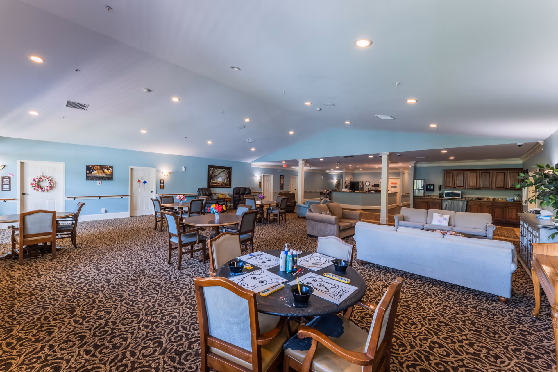 Spacious senior living common area with multiple round tables and chairs, some tables set up with coloring sheets and art supplies. Comfortable sofas and armchairs are arranged near a kitchenette area with wooden cabinets. The room has a patterned carpet, light blue walls, and recessed ceiling lights.