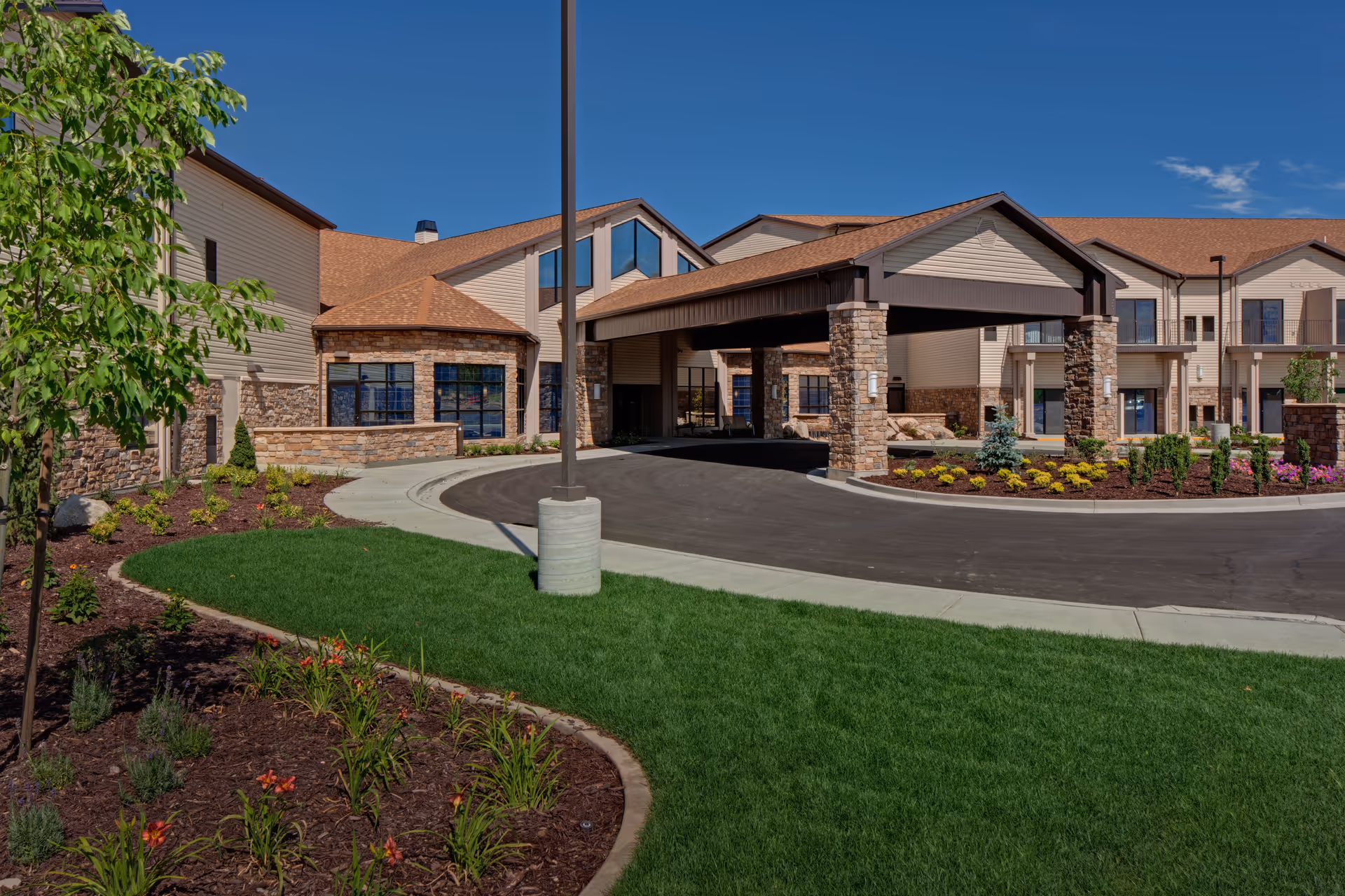 Exterior view of a senior living facility with a covered entrance supported by stone pillars, well-maintained landscaping including green grass, flowers, and shrubs, under a clear blue sky.