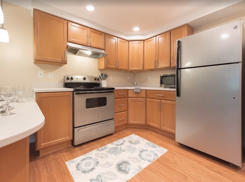 A modern kitchen with wooden cabinets, a stainless steel refrigerator, an electric stove with an overhead vent, a microwave on the countertop, and a floral patterned rug on the wooden floor. There are also some wine glasses on the left side counter.